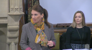 Trudy at the debate, speaking in a grey jacket with a yellow floral scarf and her hair in a ponytail